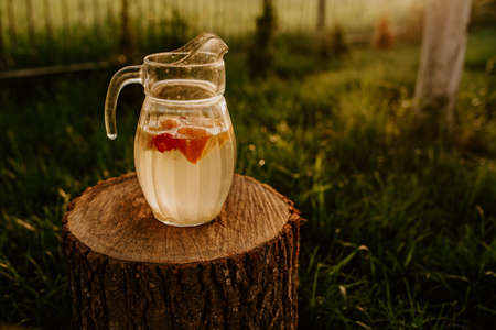 A glass jug with lemonade and fruit inside is standing on a wooden stump. at sunset on a background of green grass. strawberries, orange, lemon and mandarin.の写真素材