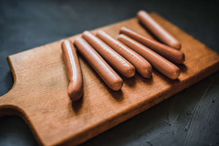 Boiled fried sausages sausages lie on a wooden kitchen board scratched against a dark concrete background.の写真素材