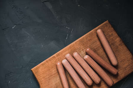 Boiled fried sausages sausages lie on a wooden kitchen board scratched against a dark concrete background.の写真素材
