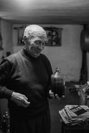 old gray-haired man with deep wrinkles drinks home-made red wine from a glass bottle in the dark basement of the house.の写真素材