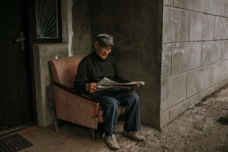A pensive old man in glasses with gray hair is sitting on an old armchair and reading a newspaper near the house. wrinkles. wisdom. against a dark gray texture wall. camouflage cap. in a knitted sweatの写真素材