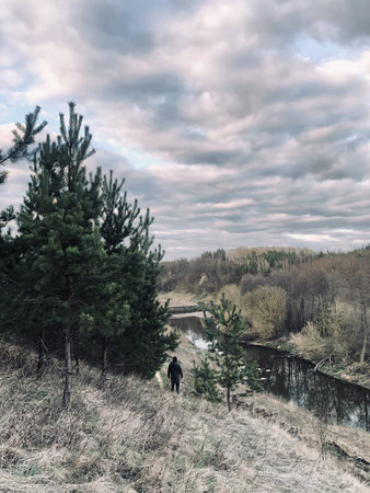 A young man, a guy of an athletic build, stands in a jacket and a hood on a hill above the river....