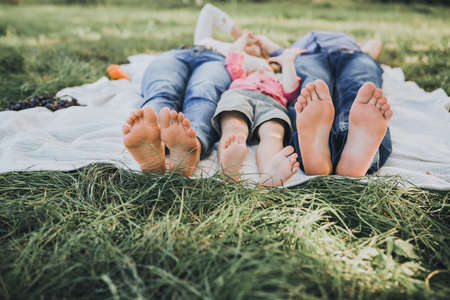 feet of a boy, mom and dad. lie on a white bedspread. Family picnic in summer in the park on green not mowed grass.の写真素材