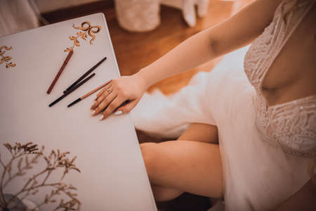 The girl sits with her legs crossed at a white cosmetic table. The bride in a white wedding dress with an open neckline. Various professional cosmetics hairbrush female accessories laid out on the tabの写真素材