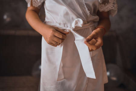 Young thin woman in pink silk robe unties the belt with her hands. bride in morning clothesの写真素材