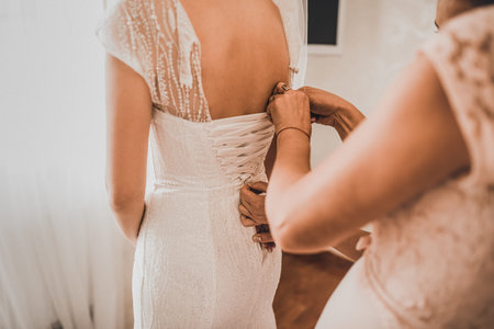 The back of the bride in a wedding dress. Moms and womans hands help fasten the clasps at the back.
