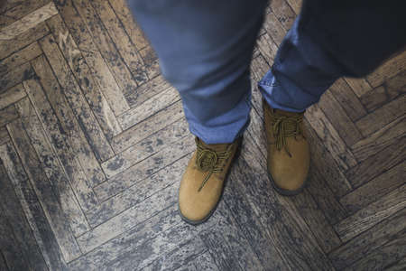 mens legs are dressed in blue denim pants and work in brown rough winter autumn shoes. Shabby old vintage original light beige wooden striped floor background.の写真素材