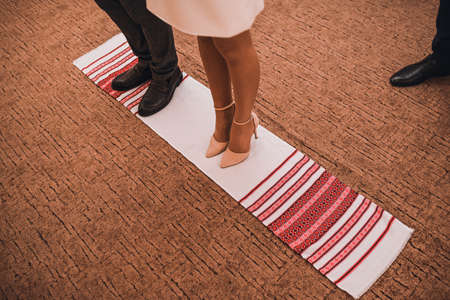 Legs of a man and a woman in classic shoes stand on a national Ukrainian towel. Black trousers and mens shoes. Womens delicate elegant beautiful pink light high-heeled shoes.の写真素材