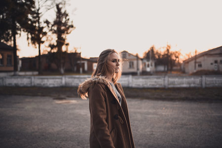 beautiful young blonde model girl. White pants. gray knitted sweater. black boots. . wooden pendant on the neck in the form of a horse. in a brown coat posing. On the Sunset. Portrait. near the treesの写真素材