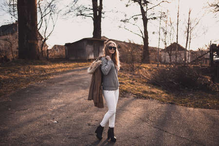 beautiful young blonde model girl. White pants. gray knitted sweater. black boots. black sunglasses. wooden pendant on the neck in the form of a horse. in a brown coat posing. On the Sunset. Portraitの写真素材