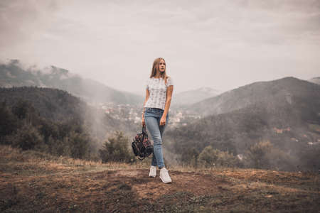 Young blonde woman standing alone with backpack on wild forest mountains at background. European girl wear tight jeans and white t-shirt sneackers. Carpathian Mountain peaks in fog scenery landscapeの写真素材
