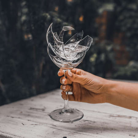 Broken crystal goblet in the hand of a woman man. Against the background of a light gray wooden shabby table and a green leafy wall. Shards folded into a glass. Ring in a glass.の写真素材