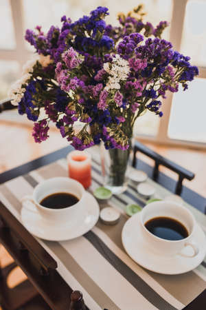 Two cups of coffee sit on a small glass table next to a bouquet of vibrant purple pink flowers in a vase. Breakfast at the hotel.の写真素材