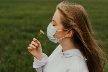 A girl in a respirator sniffs a yellow dandelion. Young blond European woman in a protective mask outdoors.の写真素材