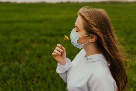 A girl in a respirator sniffs a yellow dandelion. Young blond European woman in a protective mask outdoors.の写真素材