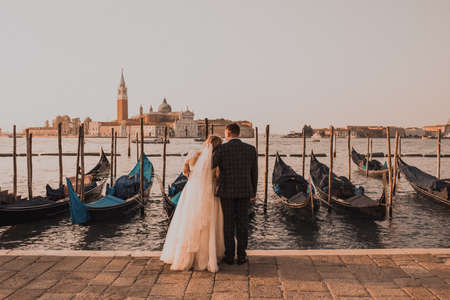 A wedding couple stands on the pier in Venice. Boats float on the water. Early morning sunrise warm summer weather. Bride in white dress groom in business black classic suit in Italy.の写真素材