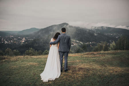 bride in a wedding dress stands her back groom in a classic suit looking at the forest in the mountains in the fogの写真素材
