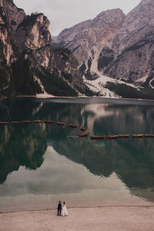 The wedding couple stands on shore of huge wide transparent turquoise aquamarine green lake in middle of high stone mountains.Brown boats float on the water.In the evening after sunset.Lago di Braiesの写真素材