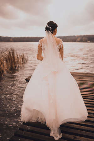 A young woman bride in white wedding dress red curly hair stands on wooden brown pier in middle of blue azure lake.の写真素材