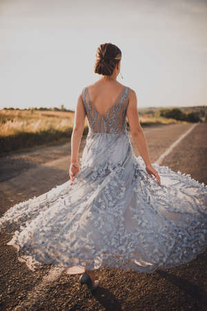 a young girl walks along an asphalt road in a gray blue long dress at sunset. back and arms open. Near the road tall yellow grass grows. The background is blurred.の写真素材