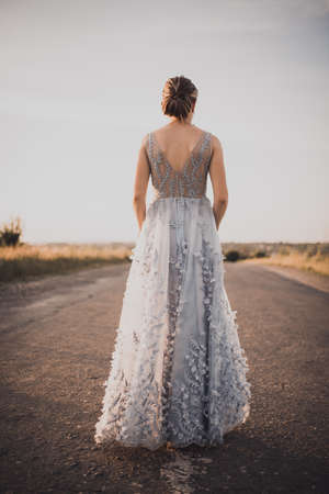 a young girl walks along an asphalt road in a gray blue long dress at sunset. back and arms open. Near the road tall yellow grass grows. The background is blurred.の写真素材
