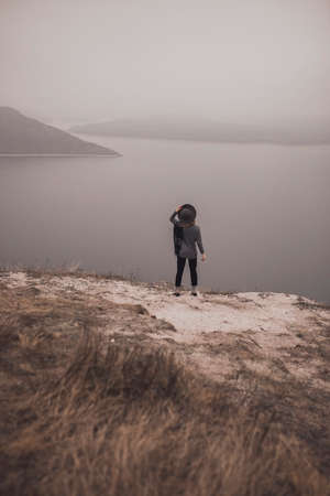 Girl traveler in a hat stands on a mountainside looks into the distance and holds a hat. the wind is blowing a scarf. Wide and smooth river. two islands away. gloomy and cold windy foggy weatherの写真素材