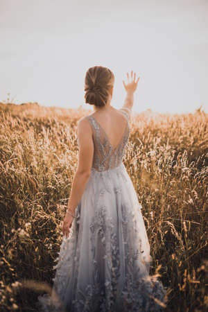 The young beautiful girl in a gray dress with an open back raised her hands up and closes the rays of the sun. against the background of blurred tall yellow grass at sunset.の写真素材