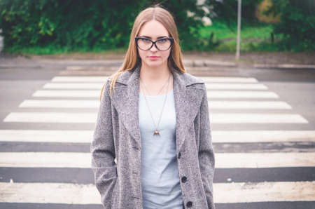 A blonde European girl in a gray coat and transparent glasses with black frames stands against the background of a pedestrian crossing.の写真素材