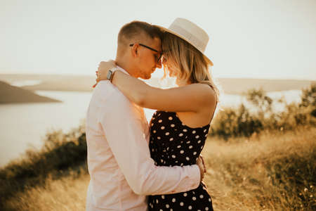 A man with a woman in a hat hug and kiss in the tall grass in the meadow. A couple of fair-haired fair-skinned people in love are resting in nature in a field at sunset.の写真素材