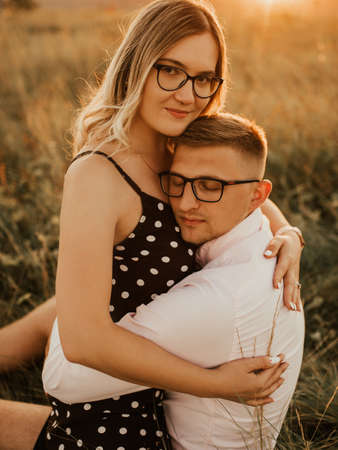 A girl in hat sits in hands guy on ground in middle of tall grass in meadow. couple of fair-haired fair-skinned people in love are resting in nature in field at sunset.の写真素材