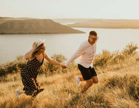 young man and woman walking in the meadow at sunset in summer near the lake. A couple of fair-haired fair-skinned people in love are resting in nature in a field at sunset.の写真素材