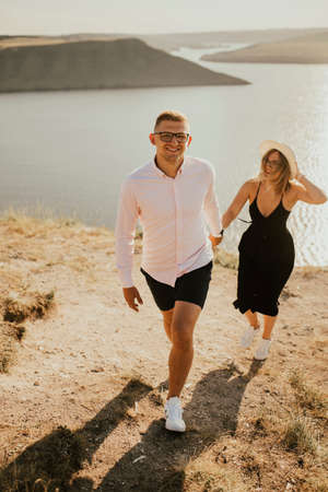 A man and a woman in love walk on a mountain above a large lake by the sea at sunset. A couple of fair-haired fair-skinned people in love are resting in nature in a fieldの写真素材