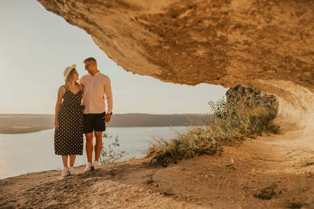 A man and a woman in a hat walk along the rocks above the cliff. A couple of fair-haired fair-skinned people in love are resting in nature in a field at sunset.の写真素材