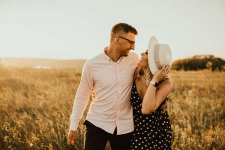 a guy with a girl in hat walking in the meadow. A couple of fair-haired fair-skinned people in love are resting in nature in a field at sunset.の写真素材
