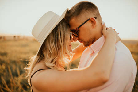 A man with a woman in a hat hug and kiss in the tall grass in the meadow. A couple of fair-haired fair-skinned people in love are resting in nature in a field at sunset.の写真素材