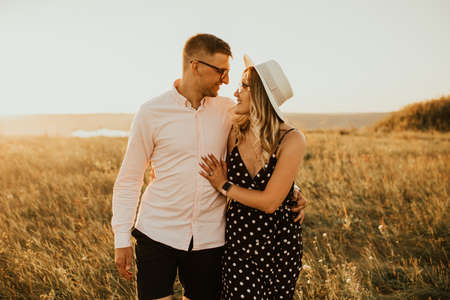 a guy with a girl in hat walking in the meadow. A couple of fair-haired fair-skinned people in love are resting in nature in a field at sunset.の写真素材