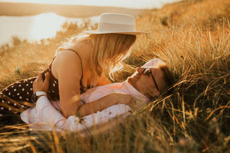 A man and a woman in a hat lie on the ground in the middle of the grass. A couple of fair-haired fair-skinned people in love are resting in nature in a field at sunset.の写真素材