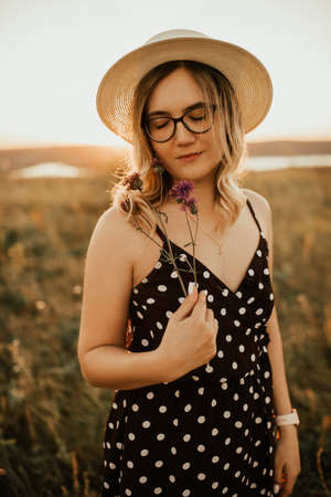 A fair-skinned blonde girl in a dress with polka dots in a hat walks through the meadow among the tall grass in the summer at sunset.の写真素材