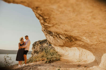 A man and a woman in a hat walk along the rocks above the cliff. A couple of fair-haired fair-skinned people in love are resting in nature in a field at sunset.の写真素材