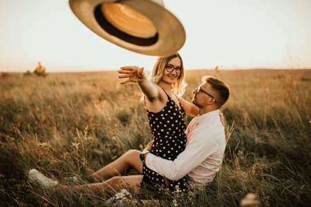 woman throws hat straight at camera. young man and woman in meadow at sunset in summer. A couple of fair-haired fair-skinned people in love are resting in natureの写真素材