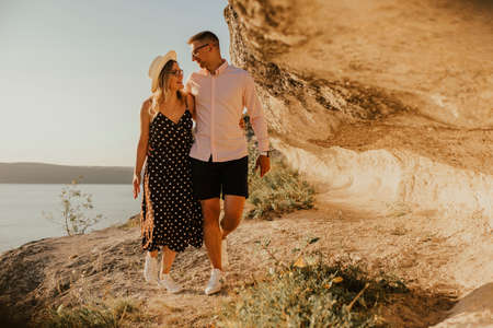 A man and a woman in a hat walk along the rocks above the cliff. A couple of fair-haired fair-skinned people in love are resting in nature in a field at sunset.の写真素材