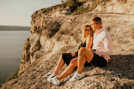 A man and a woman in a hat are sitting on a large stone rock. A couple of fair-haired fair-skinned people in love are resting in nature in a field at sunset.の写真素材