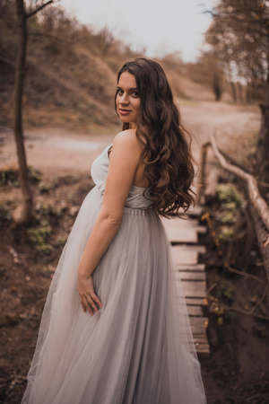 a pregnant black-haired woman with a big belly in a light long dress against a background of mountains walks along a wooden bridgeの写真素材