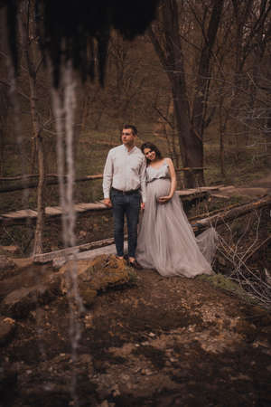 Family couple a man with a pregnant woman with a big belly in nature on the background of a waterfall. wooden bridgeの写真素材