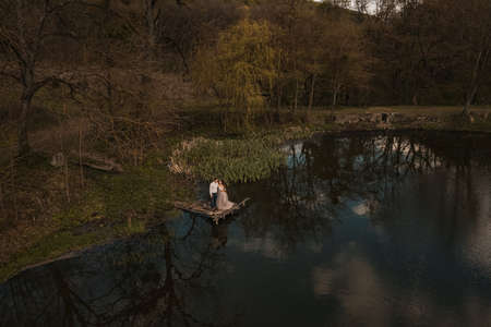 married couple a man with a pregnant woman with a big belly stand on a wooden bridge on coast lake river in nature. Aerial drone top viewの写真素材