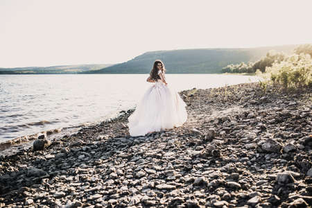 bride in wedding dress happy and laughing run against rays of setting sun in summer. bank river is strewn with stones. Bakota, Ukraineの写真素材