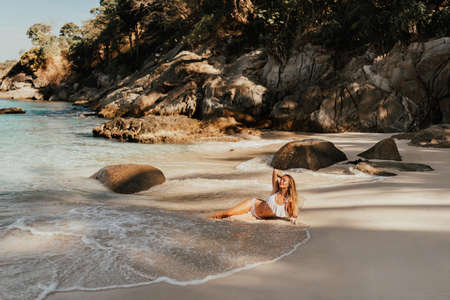 young blond European woman in white bikini swimsuit on beach lies on sand beach sea. background of large stone rocks and palm treesの写真素材