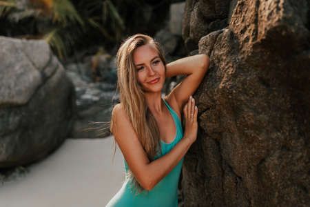 Young blonde smiling European woman in bikini swimsuit on beach against background of stone rocksの写真素材