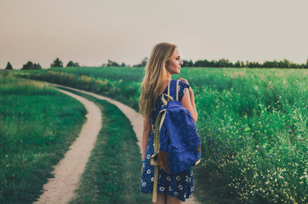 young thin blonde woman stands in dress and backpack on her shoulder in middle of green field On...