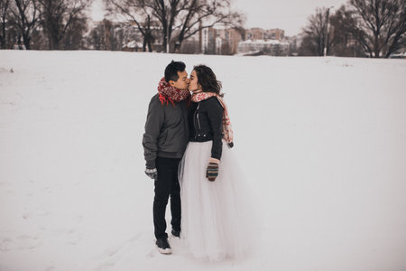 young man and woman stand kissing and hugging. bride and groom in love couple in knitted scarf and mittens in winter with snow and snowflakes. wedding dress. mexican peopleの写真素材